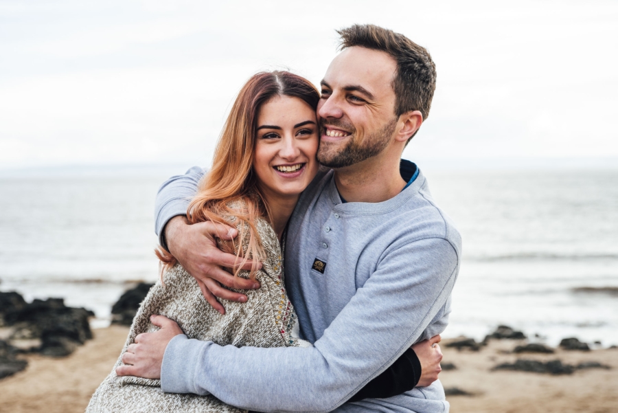 Relaxed couple photoshoot by the sea with natural smiles and warm embrace