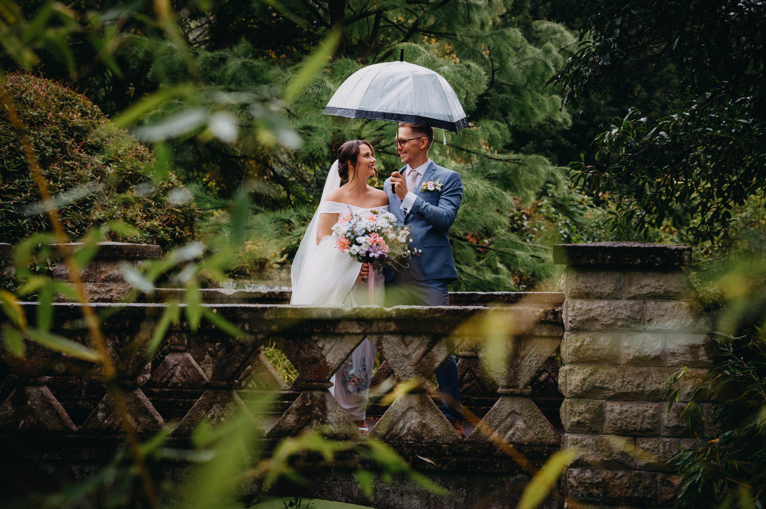 The newly married couple standing together on a stone bridge under an umbrella in the gardens near the Temple of Minerva in Bath