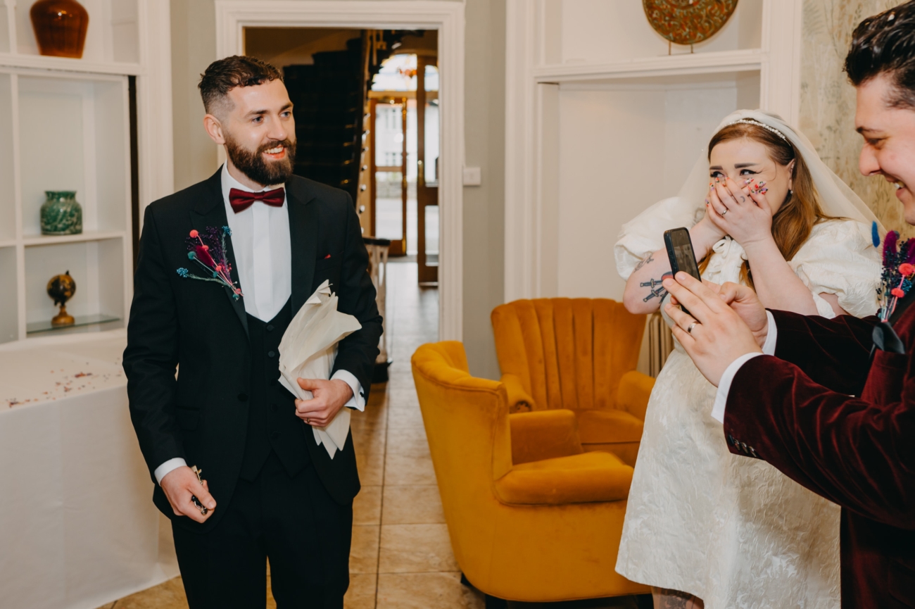 Groom photographing the best man holding printed wedding speech notes as the bride reacts with surprise
