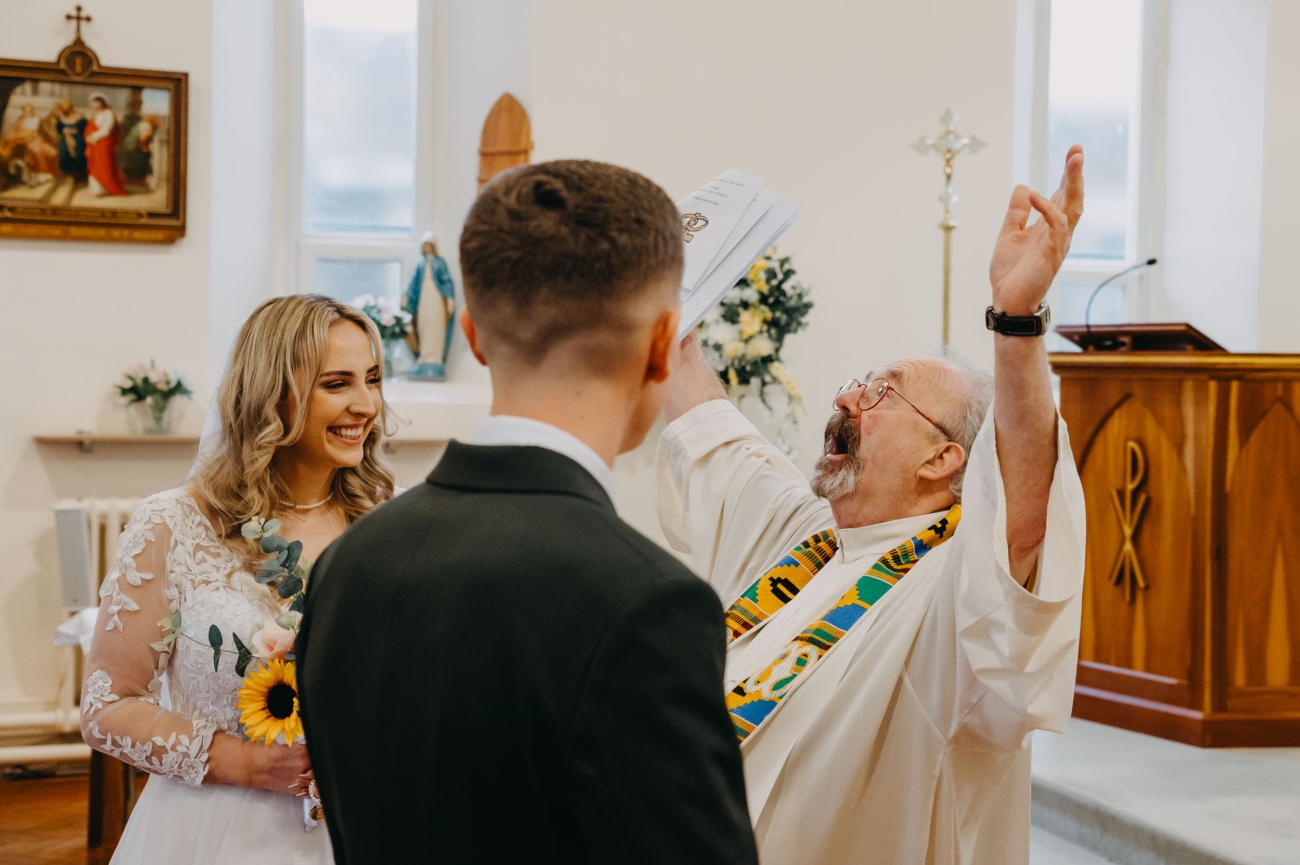 energetic wedding ceremony moment with priest raising hands and couple reacting, candid wedding photography Cardiff