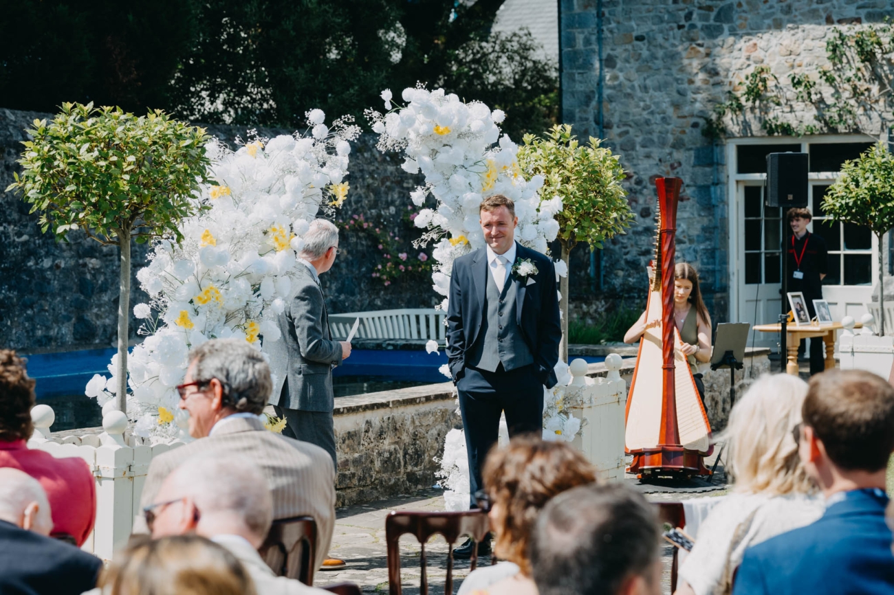 Groom smiling as he waits beside the floral arch for the outdoor ceremony to begin