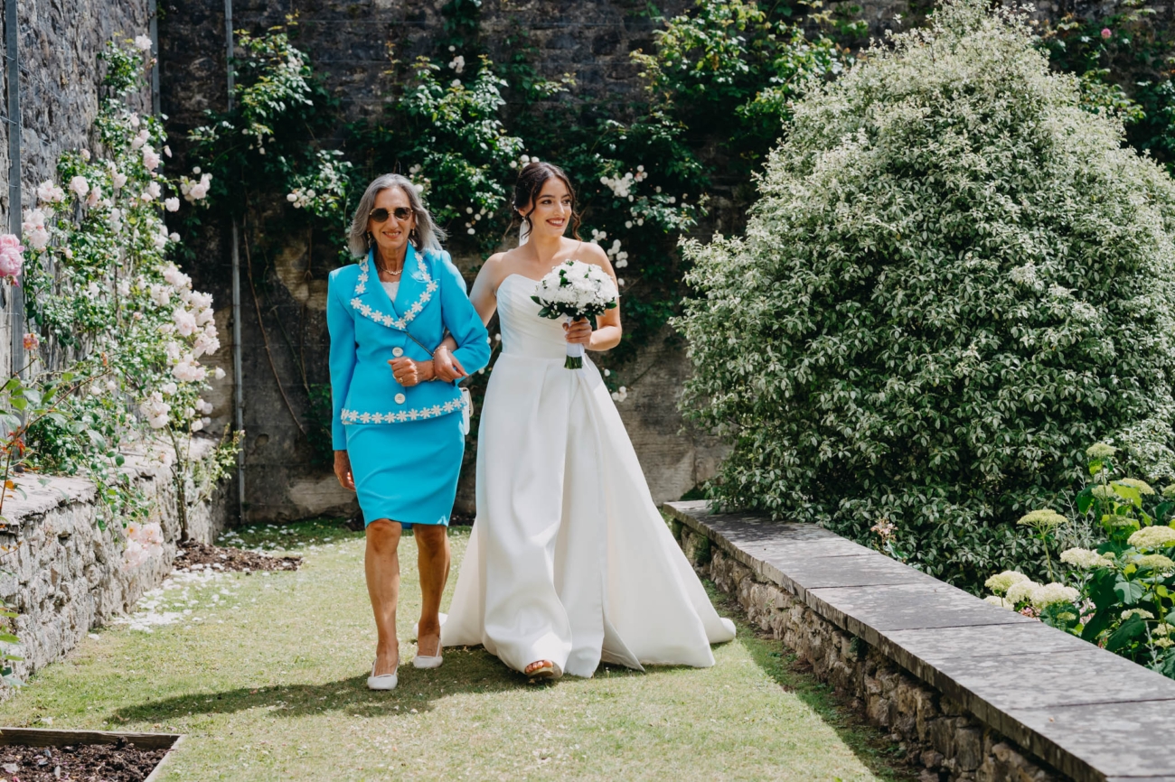 Bride walking arm in arm with a wedding guest towards the outdoor ceremony in the Italian Gardens at St Fagans