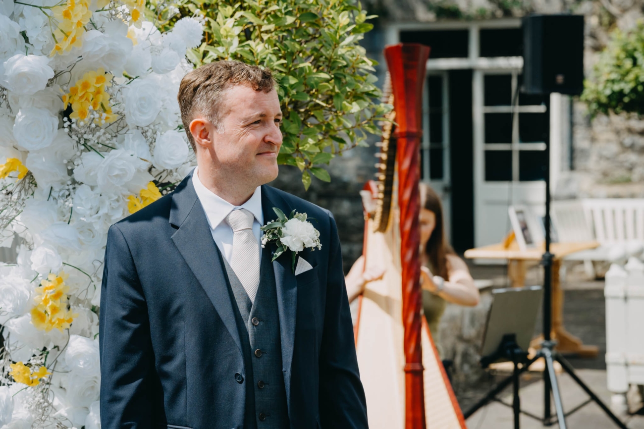 Groom smiling as he watches the bride arrive beside the floral ceremony arch