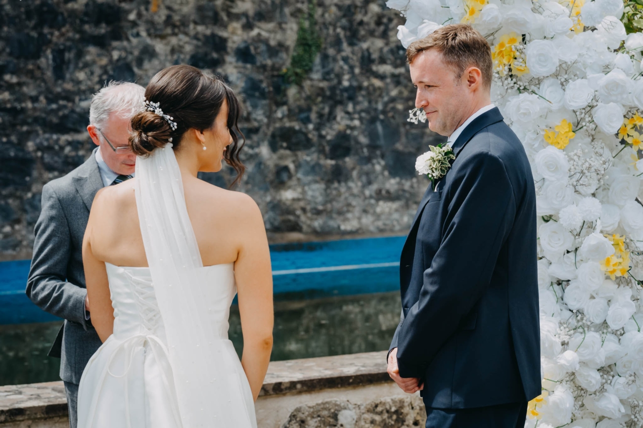Bride and groom standing face to face beside the floral arch at the beginning of their outdoor ceremony