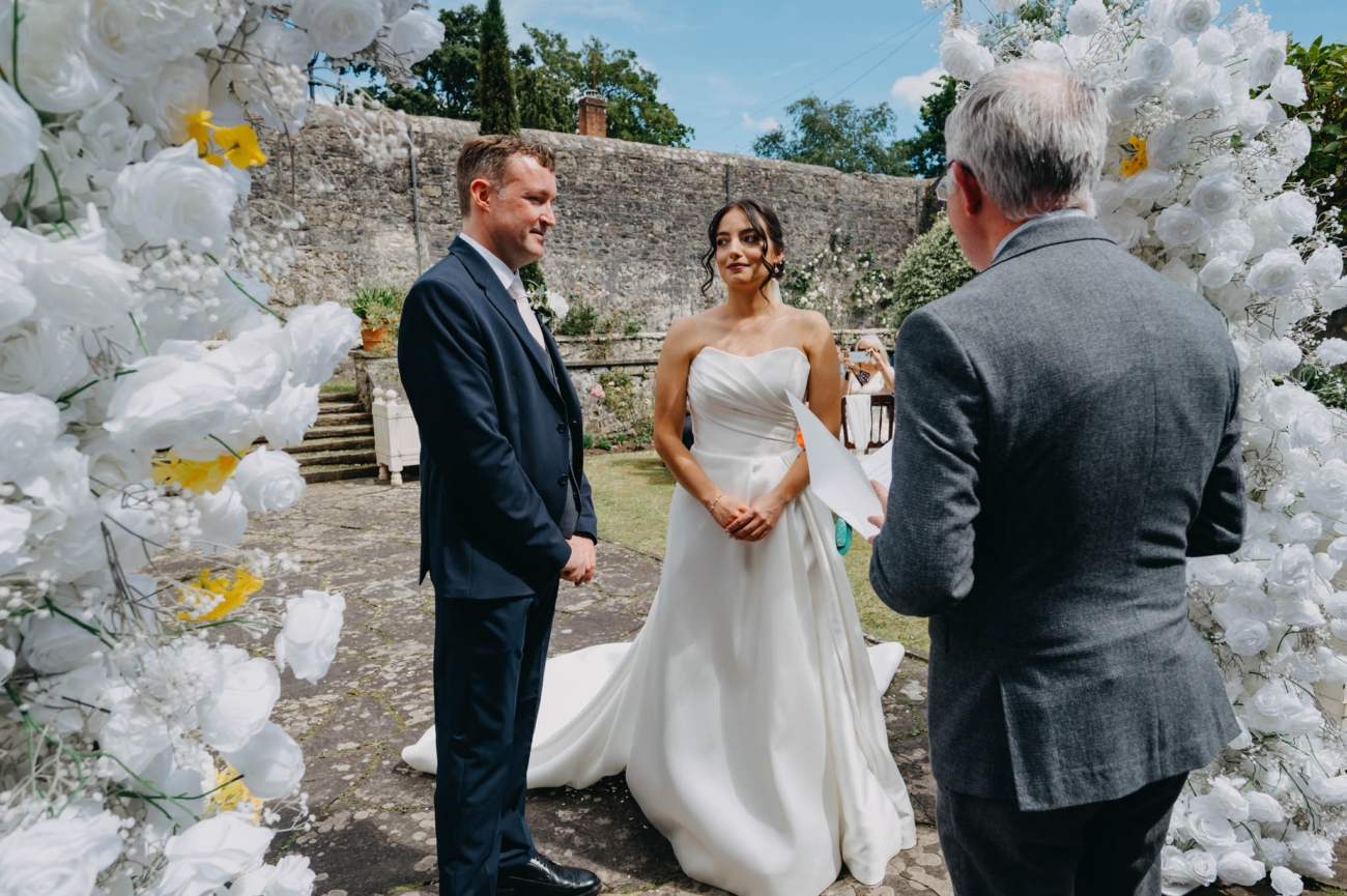 Bride and groom listening to the celebrant beneath the white floral arch in the Italian Gardens