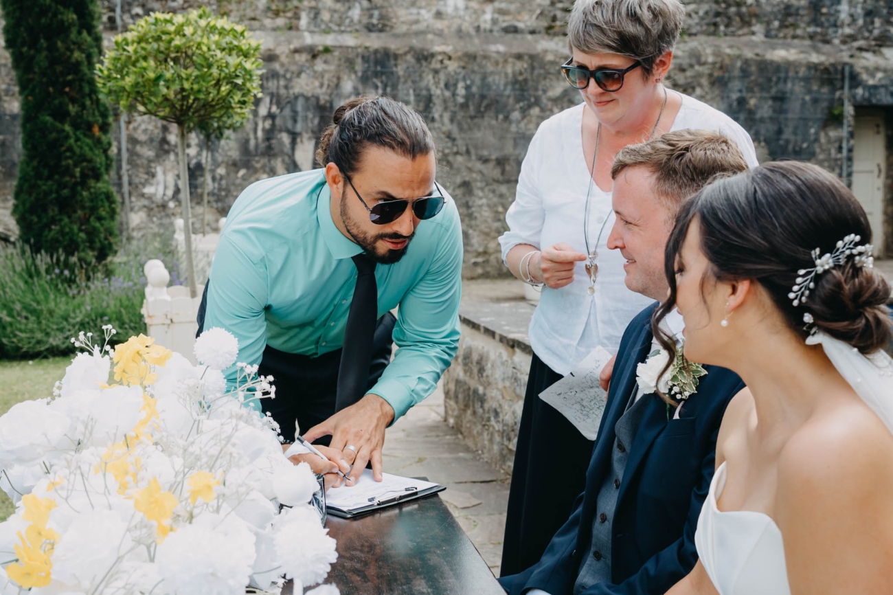Wedding guest signing the register as the bride and groom look on during the outdoor ceremony