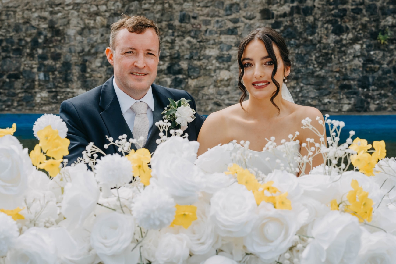Bride and groom seated together behind white and yellow flowers during their outdoor wedding ceremony