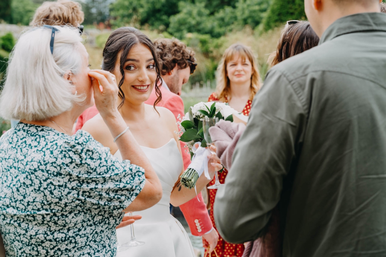 Bride smiling as she talks with wedding guests while holding her bouquet and a glass of sparkling wine
