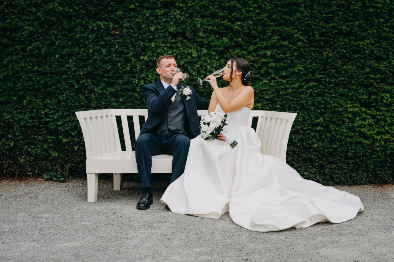 Newlyweds drinking sparkling wine while seated together on a white bench in the Italian Gardens