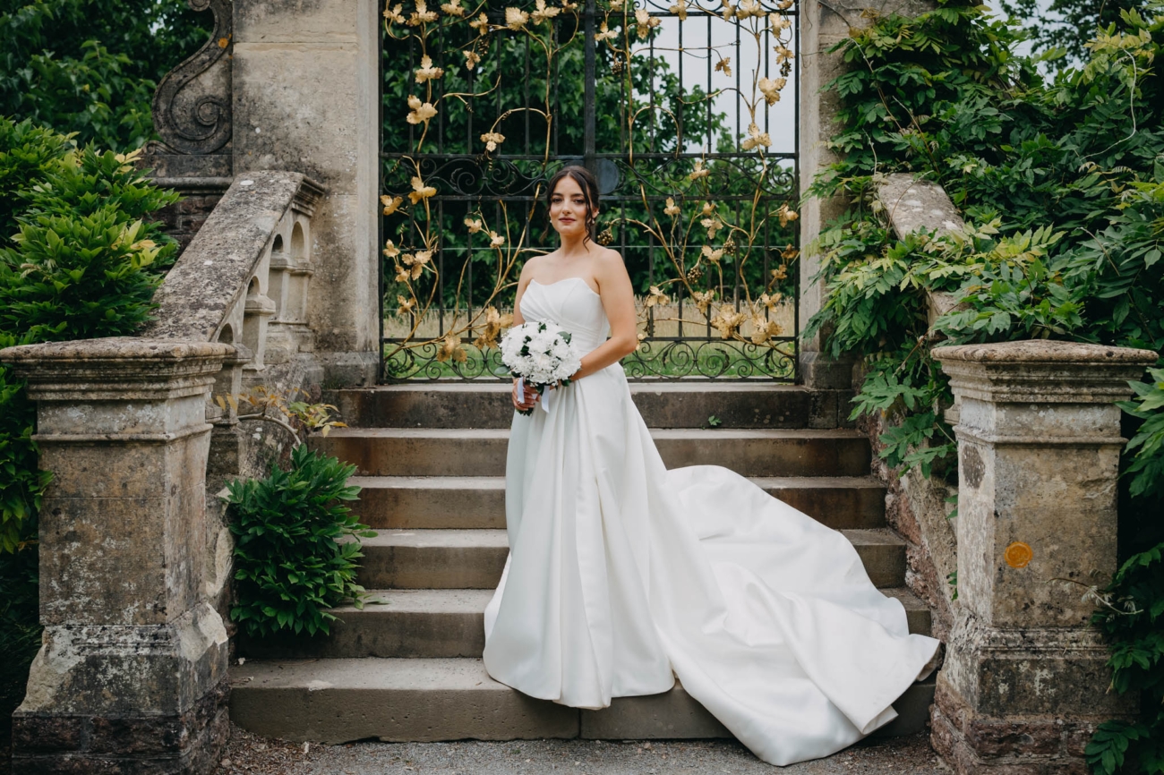 Bride holding her white bouquet on the stone steps in front of an ornate gate in the Italian Gardens