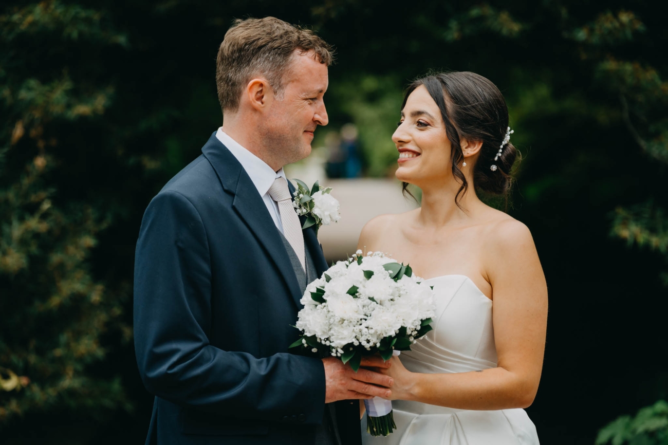 Bride and groom smiling at each other while holding a white wedding bouquet