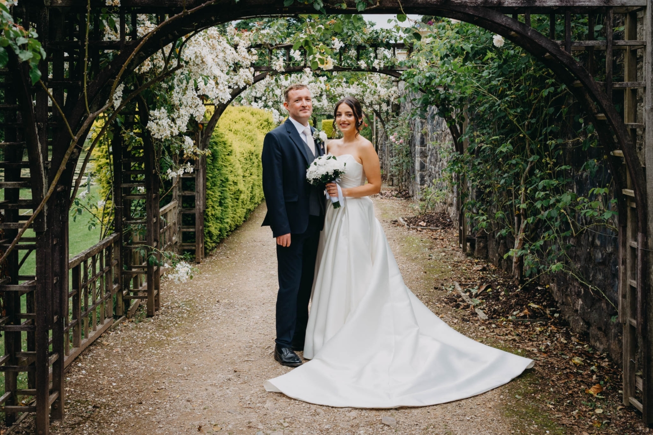 Bride and groom standing beneath a flower-covered wooden archway in the Italian Gardens at St Fagans