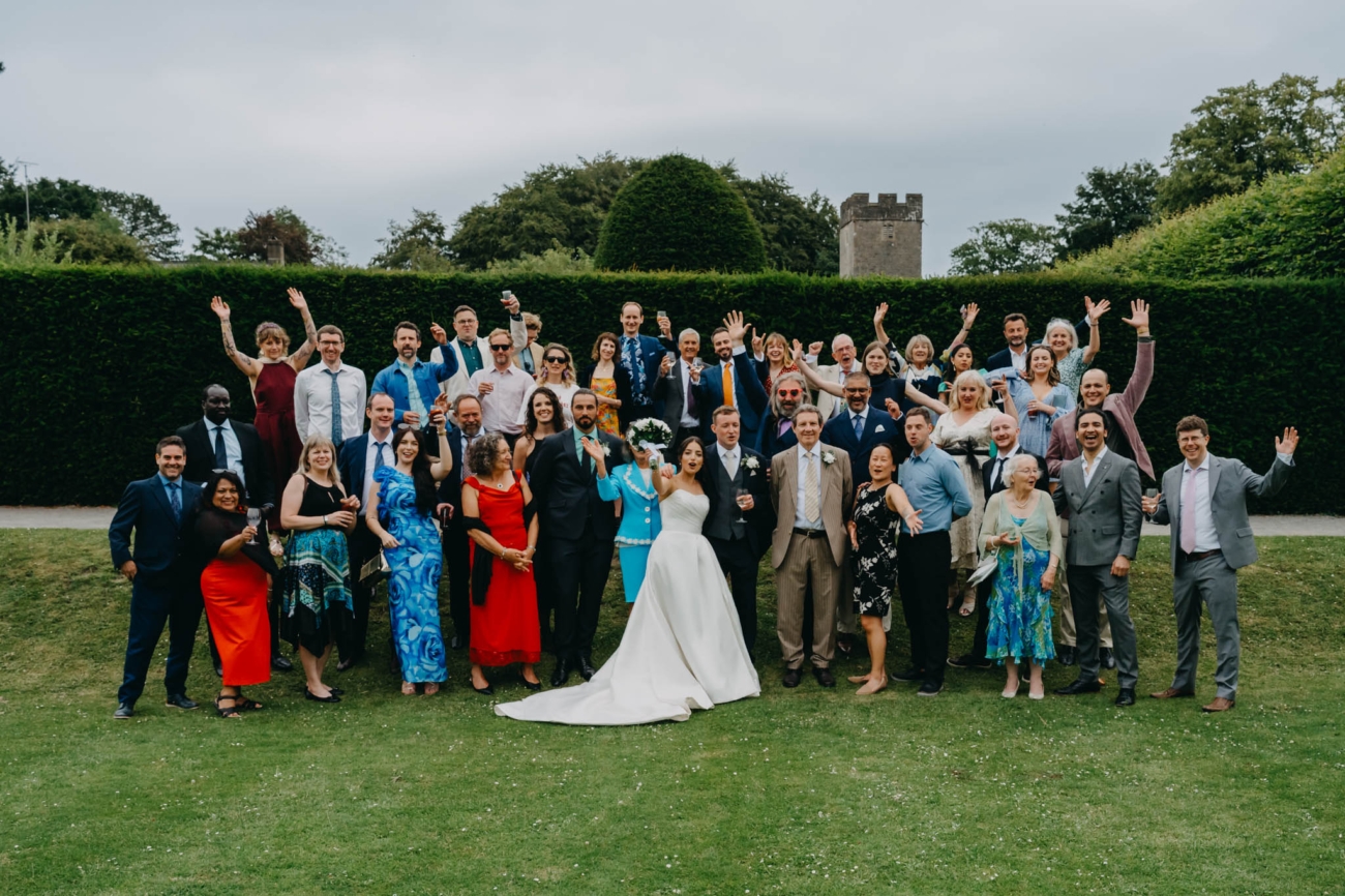 Bride, groom and wedding guests raising their hands during a large group photograph in the Italian Gardens
