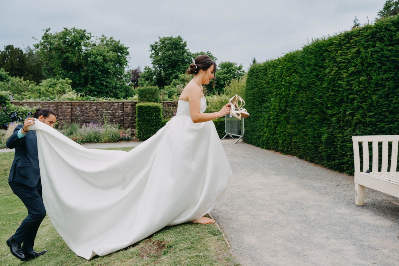 Barefoot bride carrying her shoes while a wedding guest lifts the train of her dress