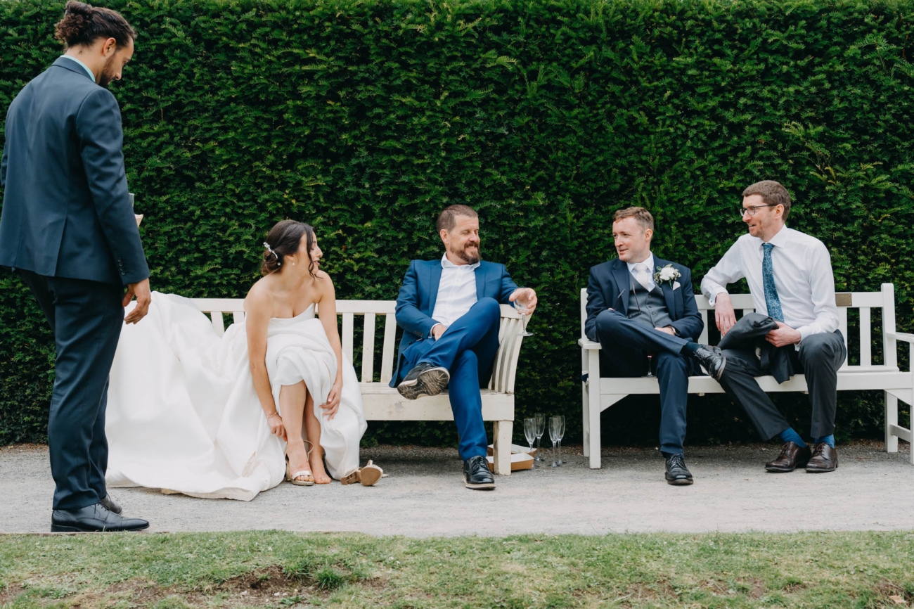 Bride fastening her shoe while the groom and wedding guests relax on white garden benches