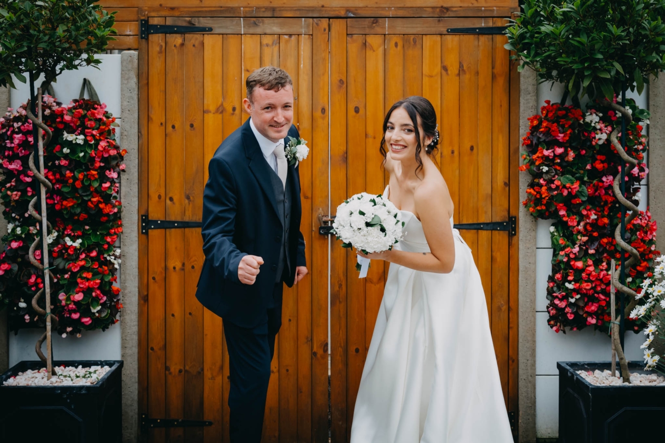 Bride and groom smiling in front of wooden doors framed by colourful hanging flowers