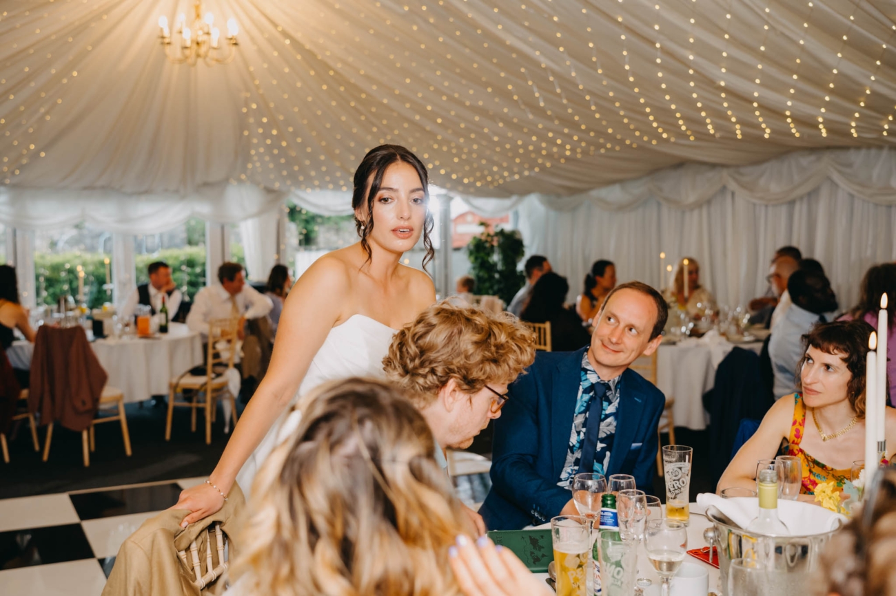 Bride chatting with wedding guests seated around a reception table inside the marquee