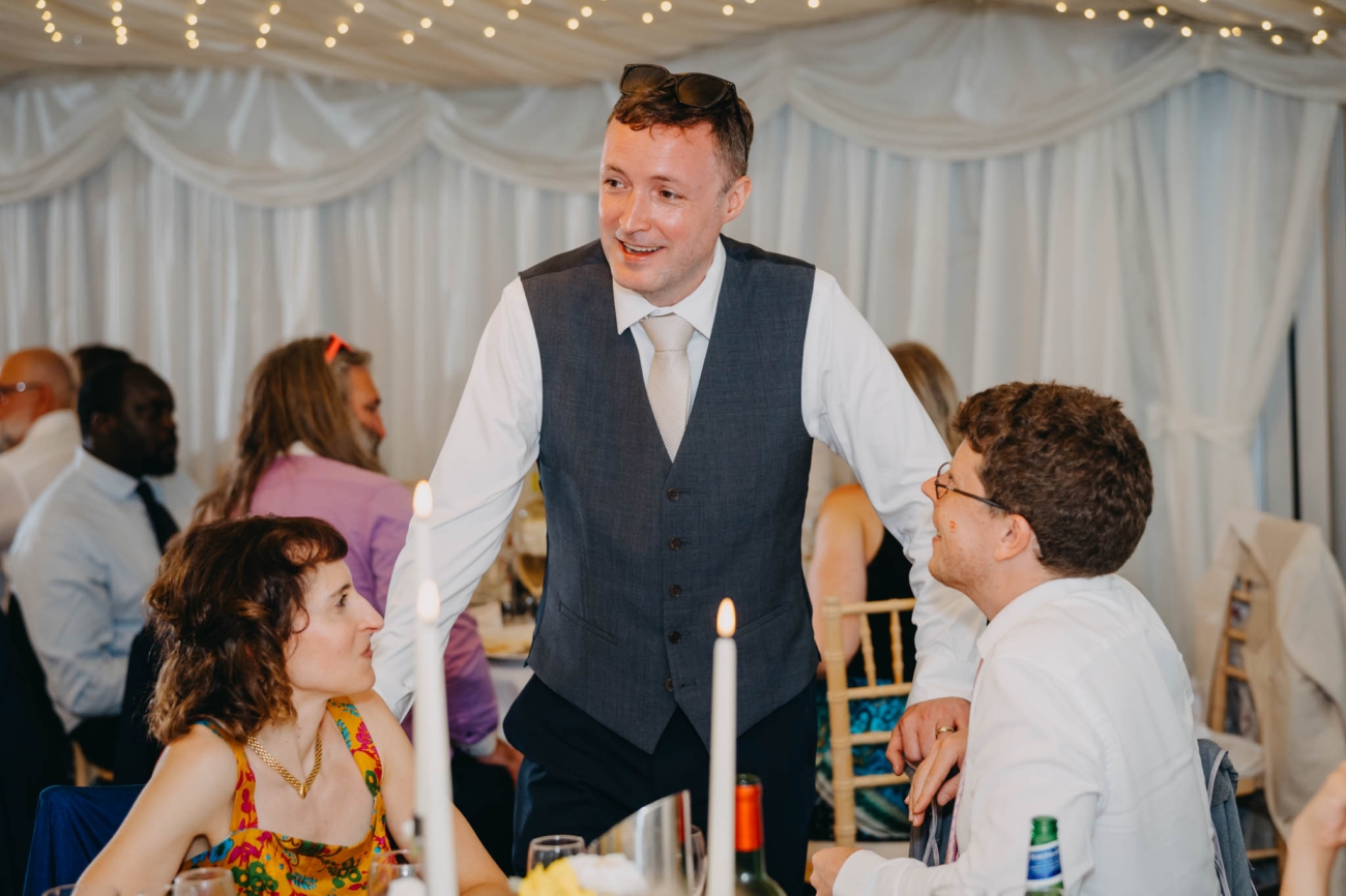 Groom standing beside a table and chatting with seated wedding guests