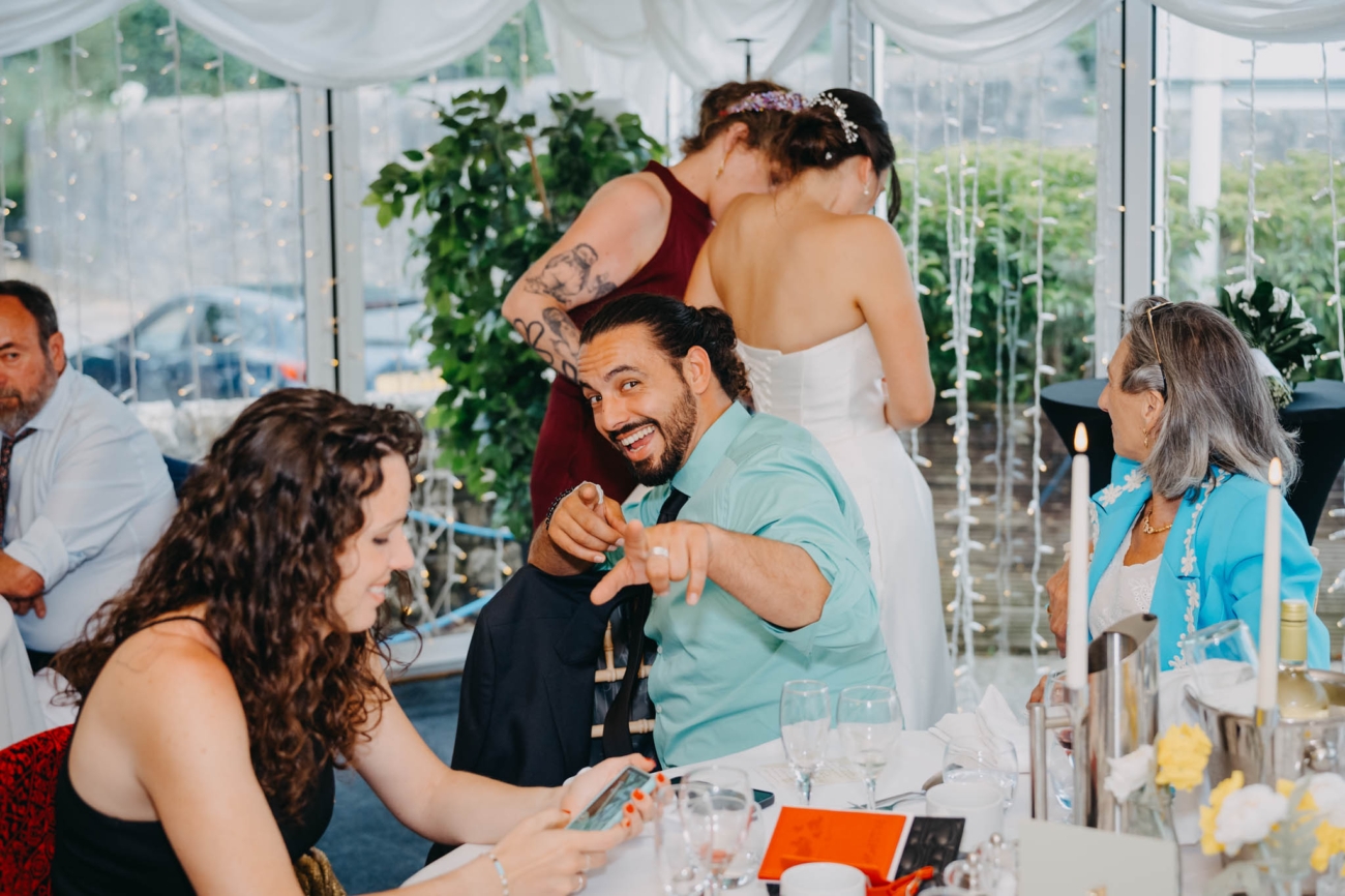 Wedding guest pointing playfully towards the camera as the bride talks with another guest behind him