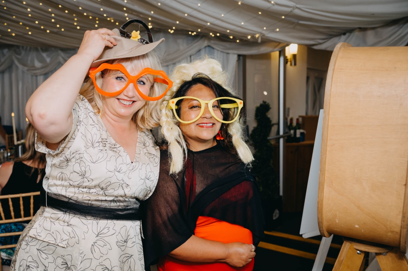 Two wedding guests posing in hats and oversized glasses beside the photo booth