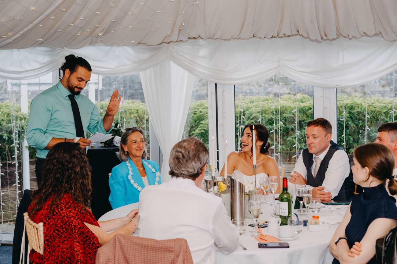 Wedding guest delivering a speech from handwritten notes as the bride laughs at the top table