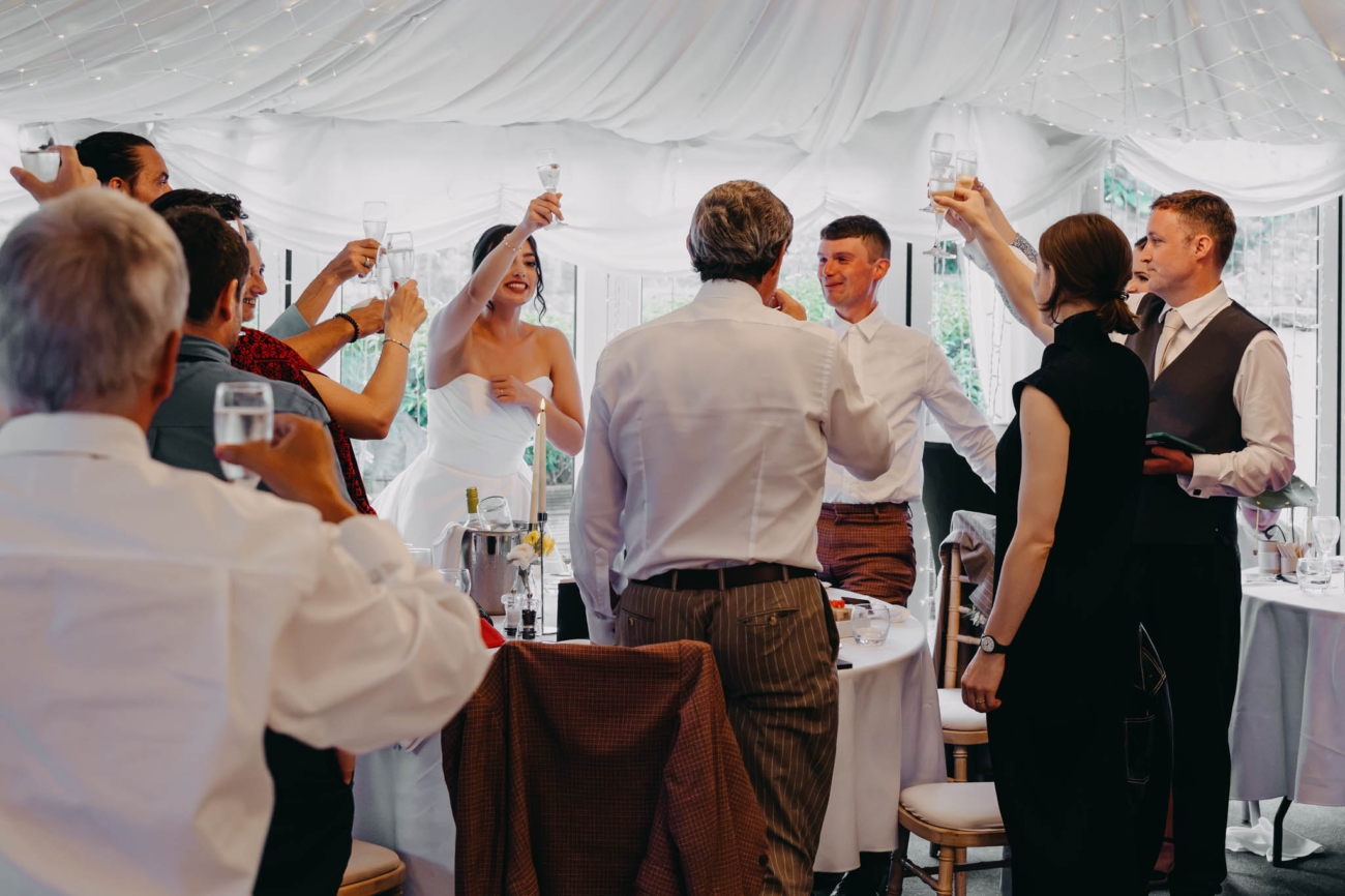 Bride, groom and wedding guests raising their glasses for a toast inside the Old Post House marquee