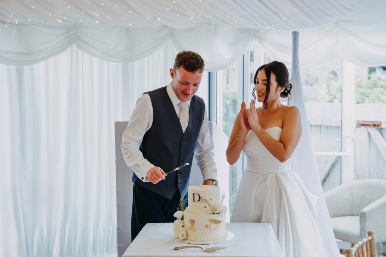 Bride clapping as the groom cuts their white and gold wedding cake