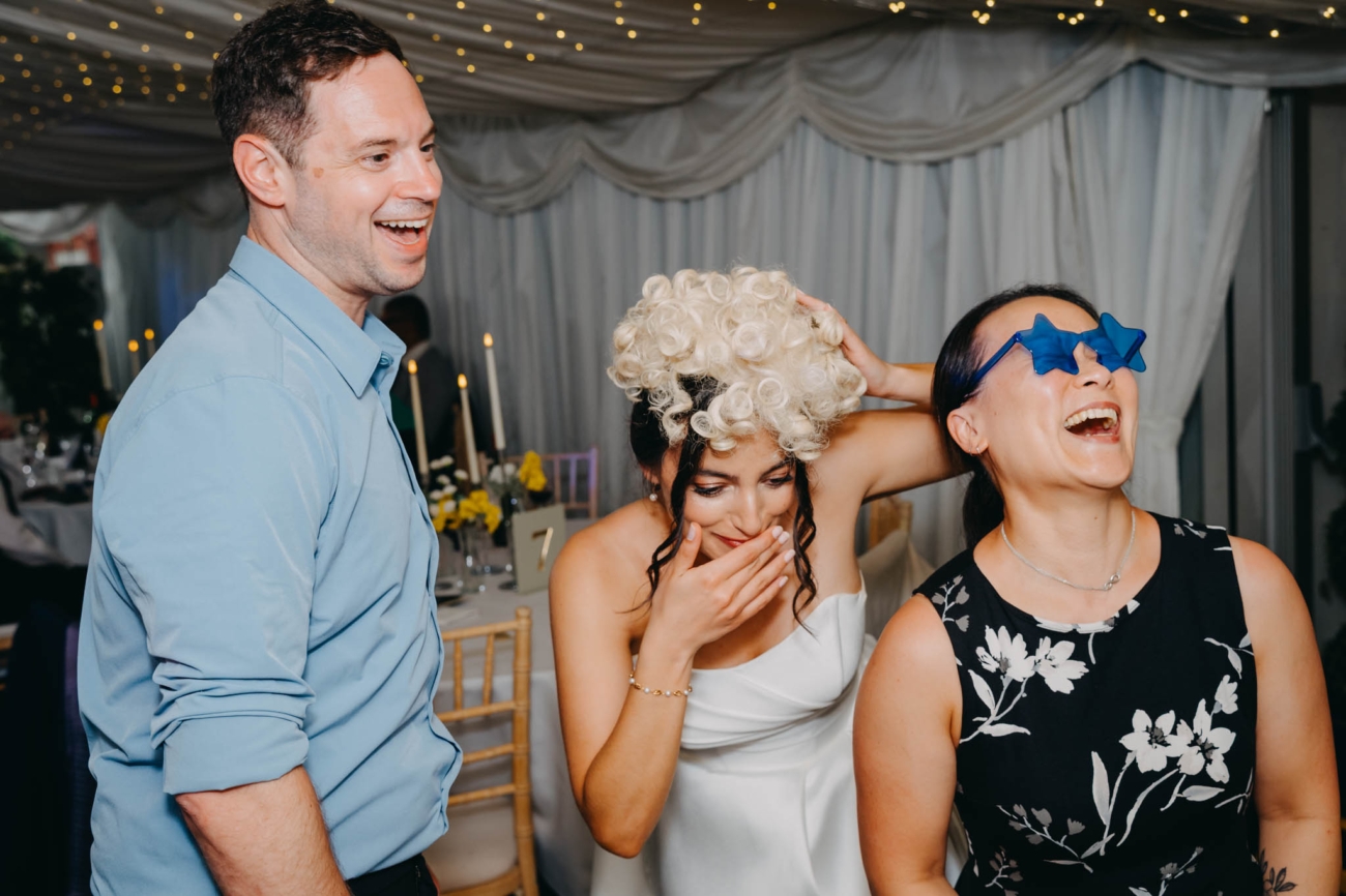 Bride wearing a curly wig beside a guest in star-shaped glasses as they laugh near the photo booth