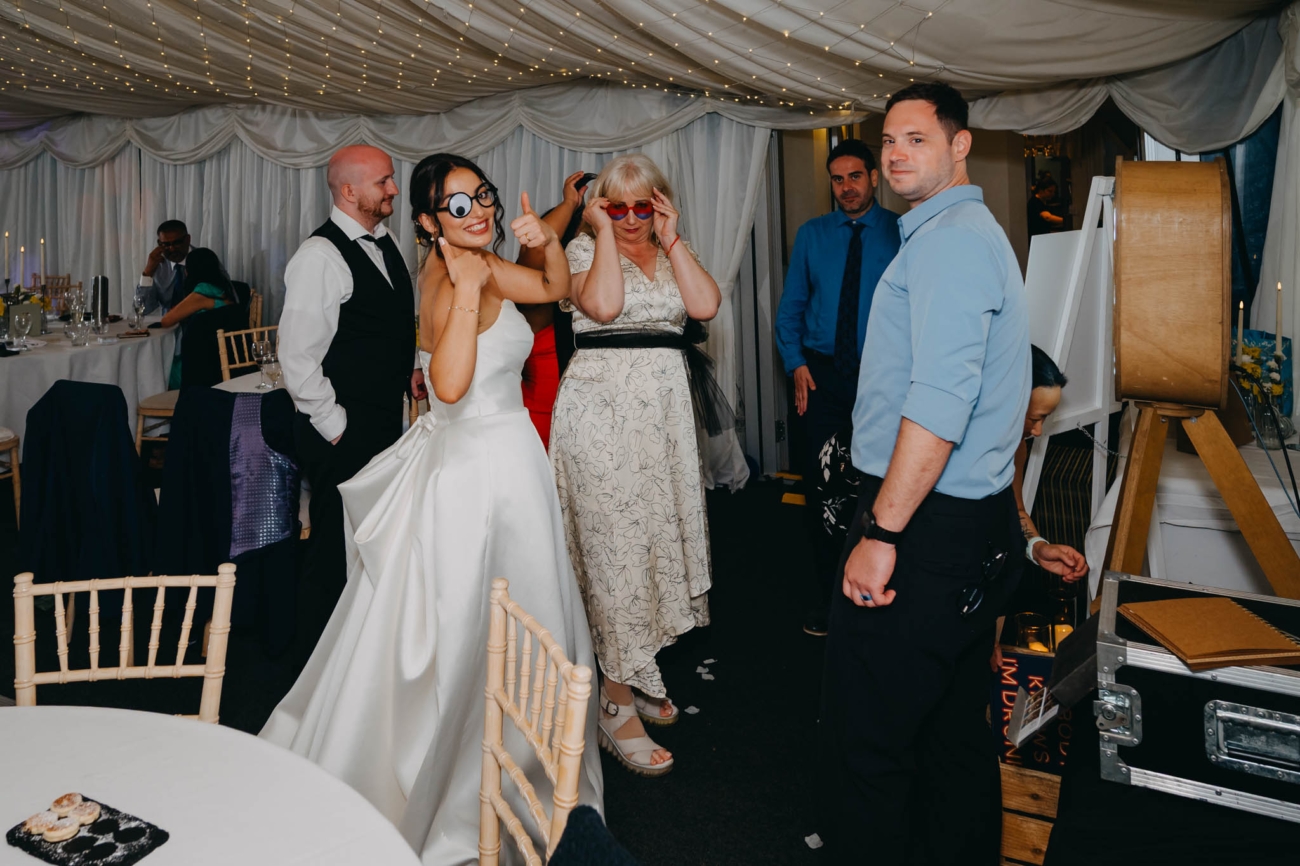 Bride and wedding guests posing in wigs and oversized glasses beside the photo booth