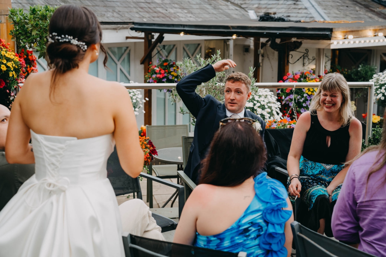 Groom gesturing animatedly as the bride and wedding guests laugh on the terrace