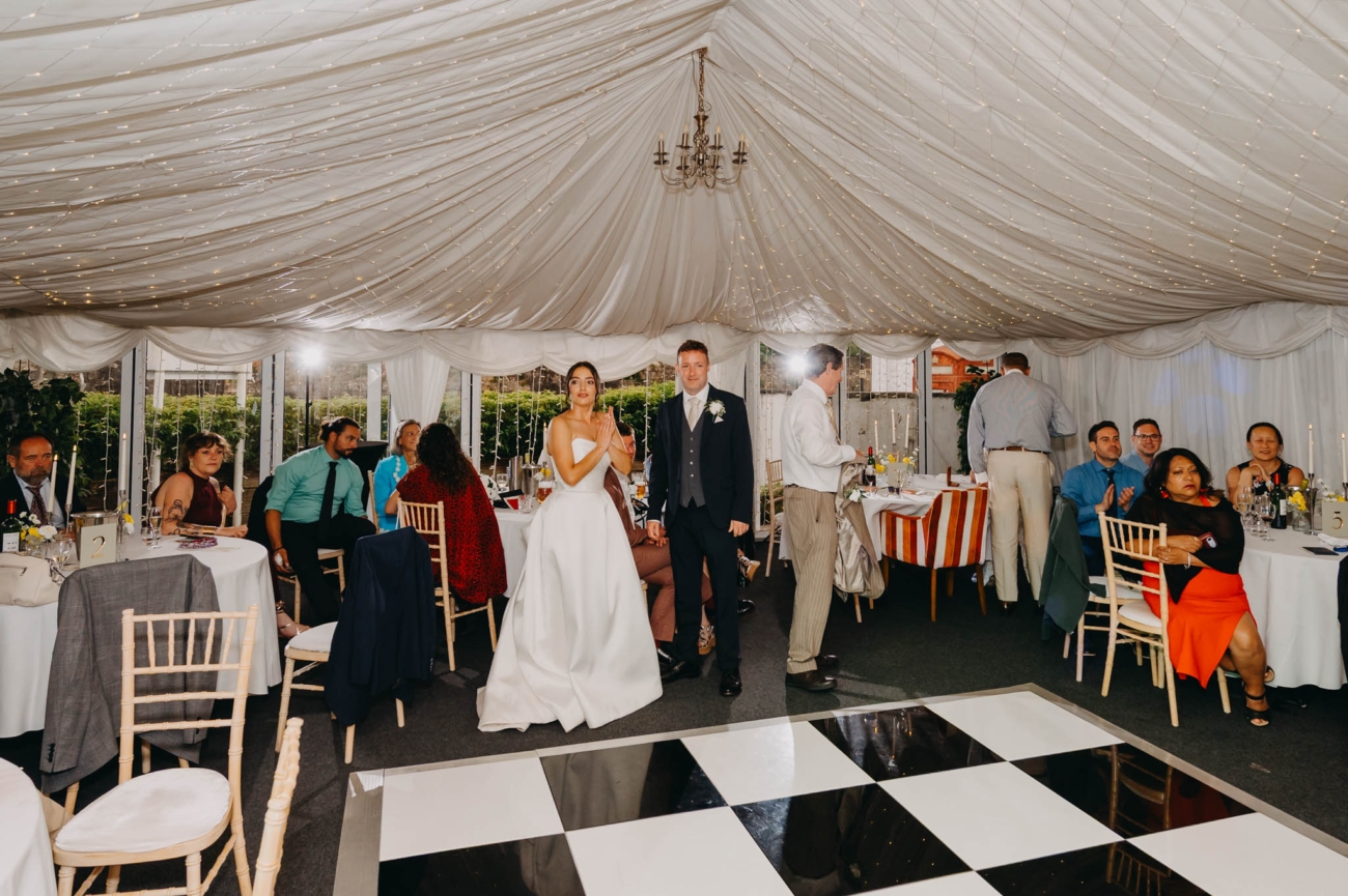 Bride and groom standing beside the dance floor as guests applaud inside the Old Post House marquee