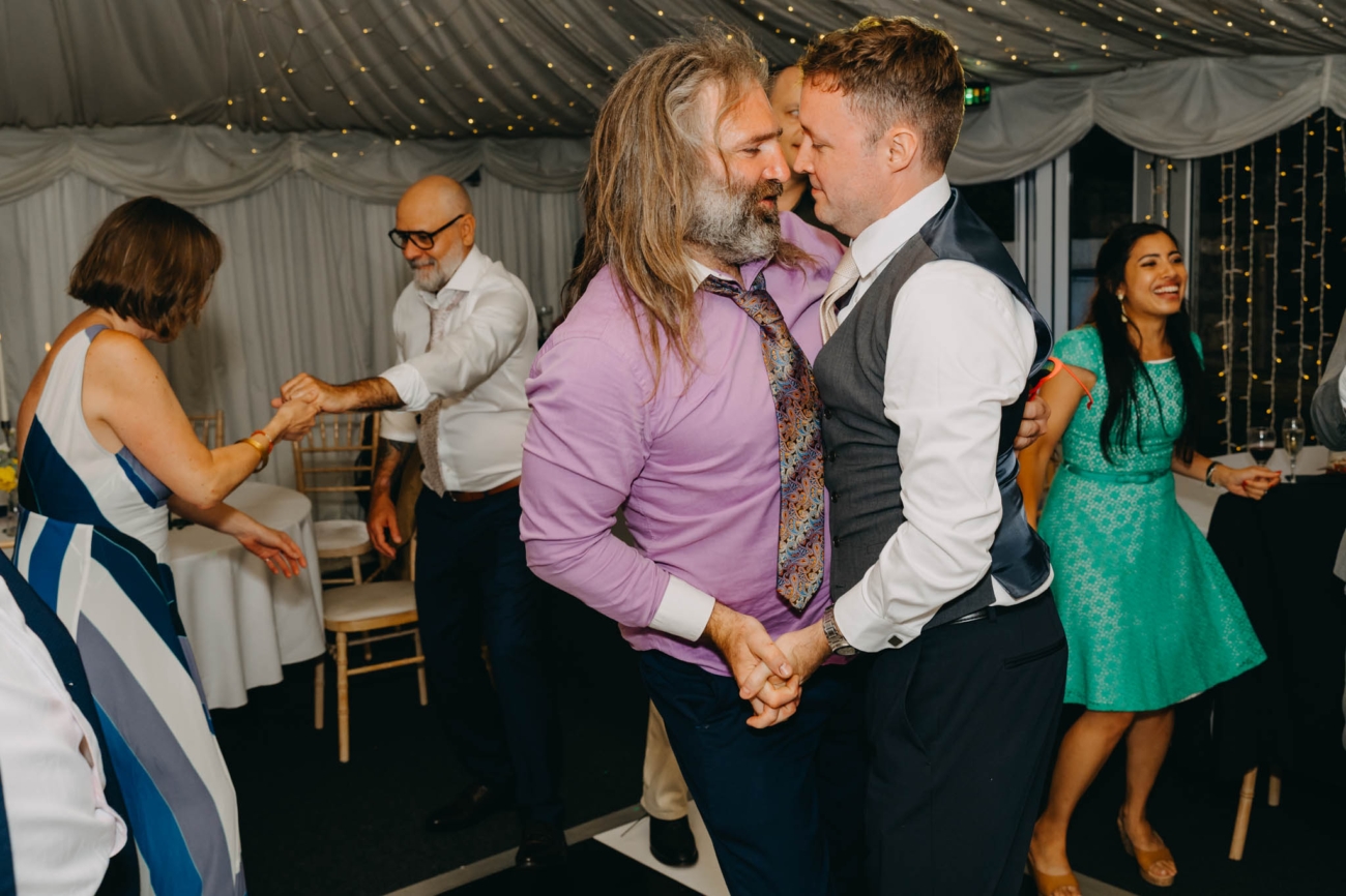 Groom dancing face to face with a long-haired wedding guest as others dance around them