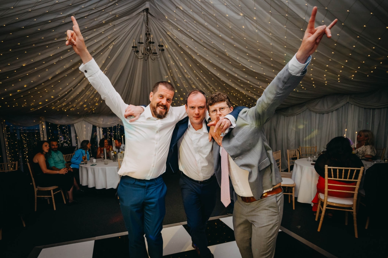 Three wedding guests dancing with their arms raised beneath the marquee lights