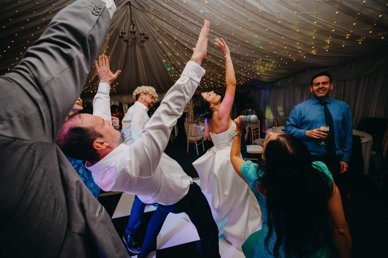 Bride and wedding guests singing into a microphone with their hands raised on the dance floor