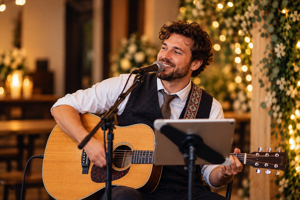 Wedding musician singing and playing acoustic guitar at a wedding reception, surrounded by warm lights and floral decorations.