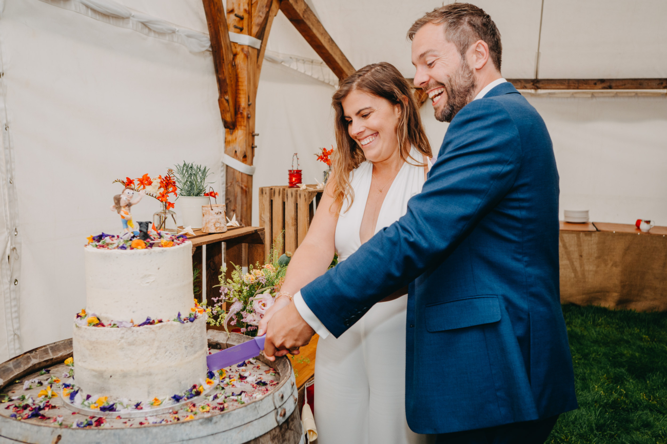 Bride and groom smiling as they cut their two-tier wedding cake decorated with colourful flowers during their reception.