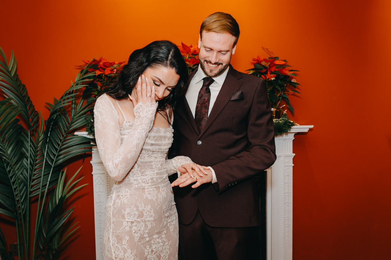 Bride covering her face with emotion while the groom smiles and holds her hand during their wedding ceremony.