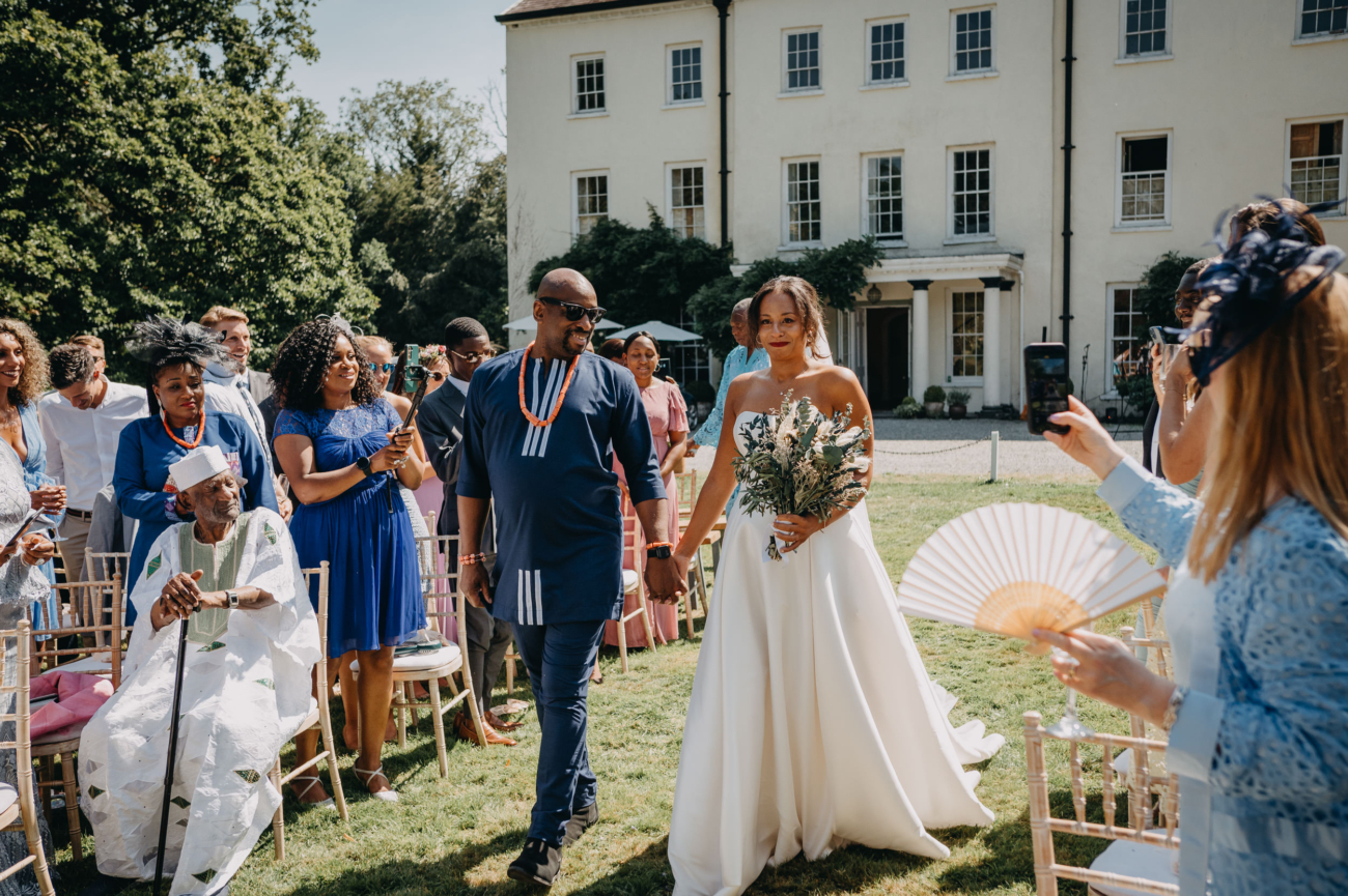 Bride walking down the aisle holding hands with a man during an outdoor wedding ceremony, surrounded by smiling guests in front of a country house.