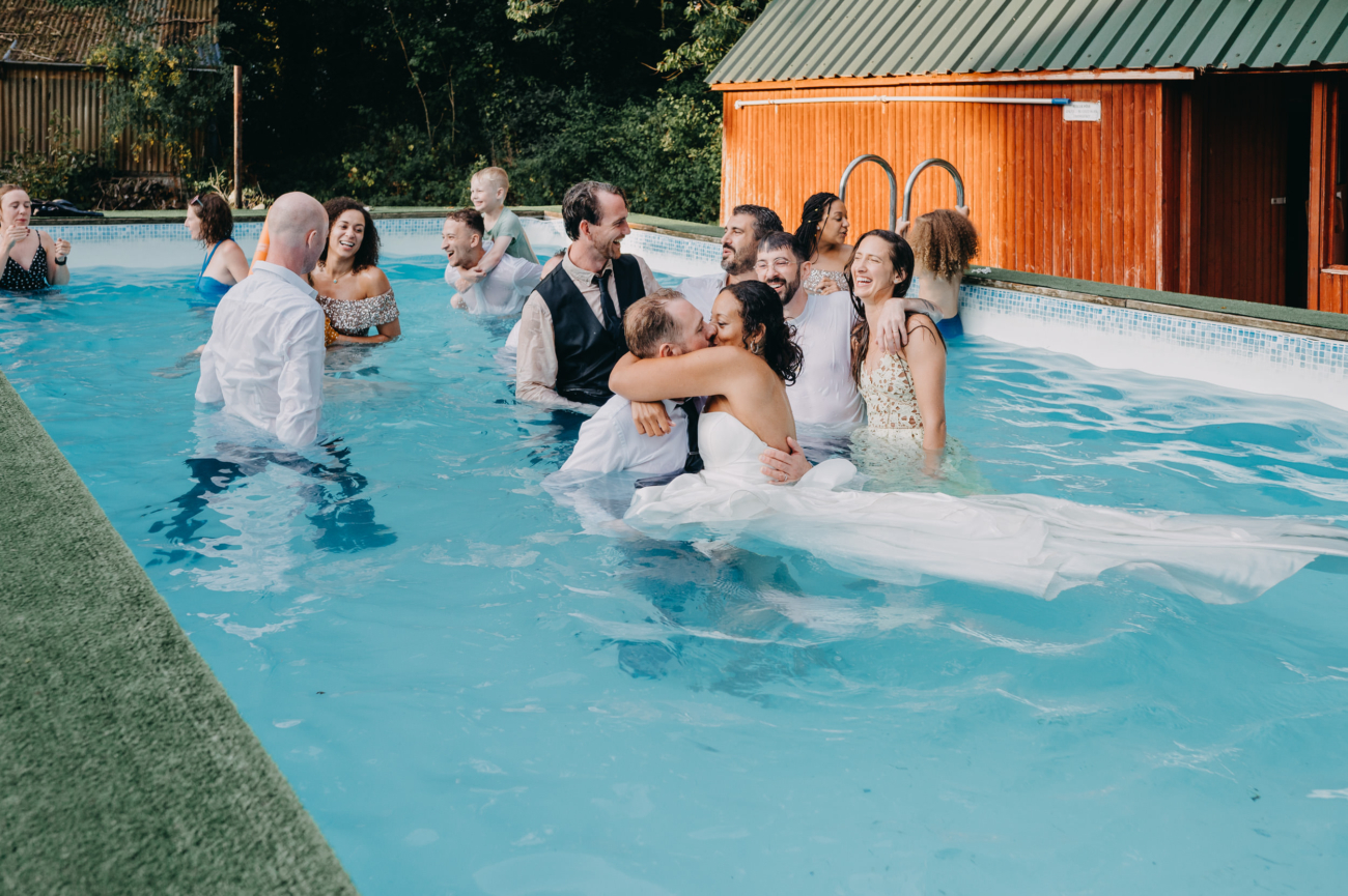 Bride and groom kissing in a swimming pool surrounded by laughing wedding guests during a spontaneous celebration.