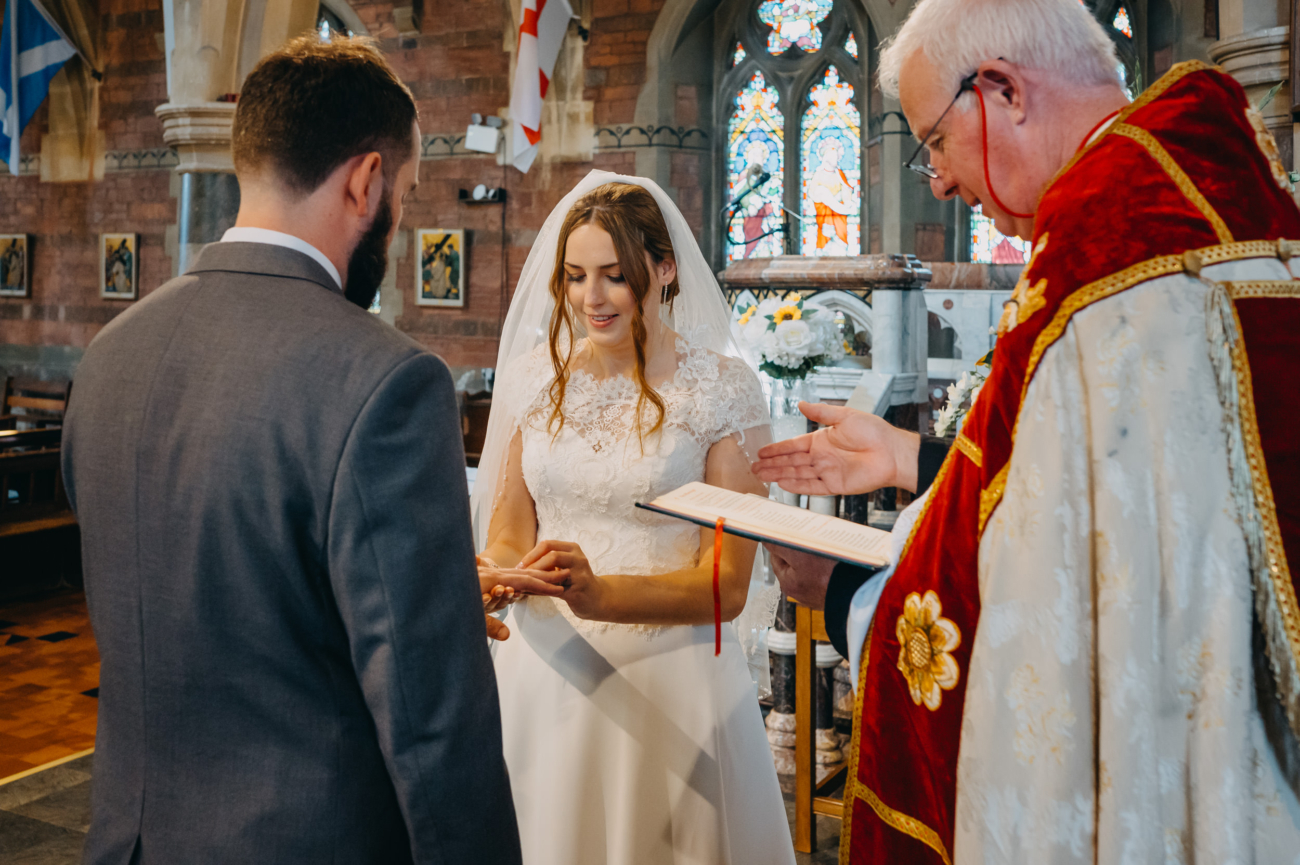 Bride placing a wedding ring on the groom's finger during a church ceremony, with the priest officiating beside them.