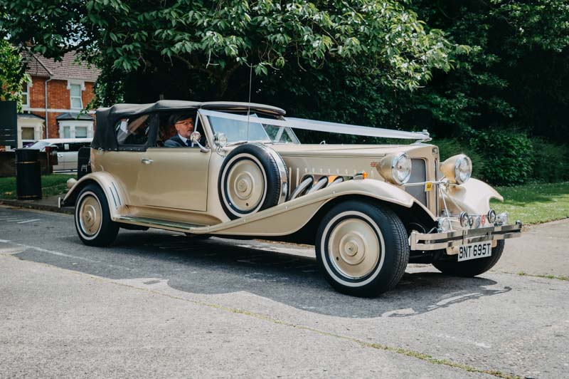 Cream vintage wedding car decorated with white ribbons, parked beside a tree-lined road