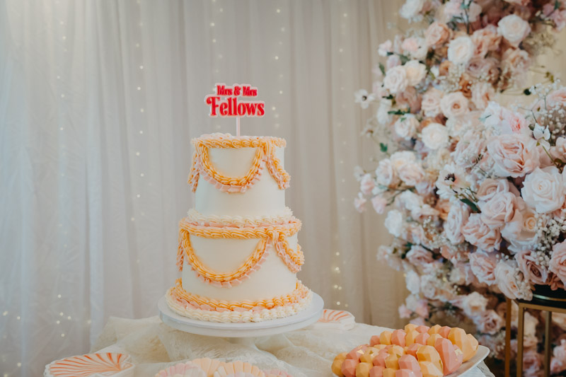 Two-tier wedding cake with orange piping beside a large arrangement of pink and white flowers