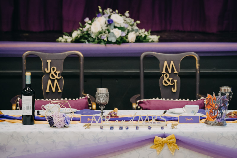 Wedding top table decorated with flowers, personalised chairs, purple ribbons and gold details